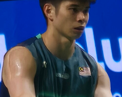Close-up portrait of Malaysian badminton player Leong Jun Hao wearing a dark green athletic jersey with the national flag emblem during a match.