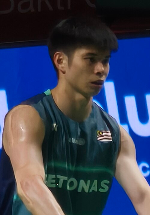 Close-up portrait of Malaysian badminton player Leong Jun Hao wearing a dark green athletic jersey with the national flag emblem during a match.