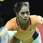K. Letshanaa lunges forward to execute a low forehand net return in her orange Malaysian national jersey during an international badminton match.