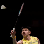 Leong Jun Hao reaches up to strike an overhead return in his yellow Malaysian national jersey during an international badminton match.