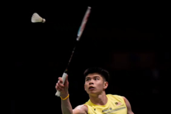 Leong Jun Hao reaches up to strike an overhead return in his yellow Malaysian national jersey during an international badminton match.