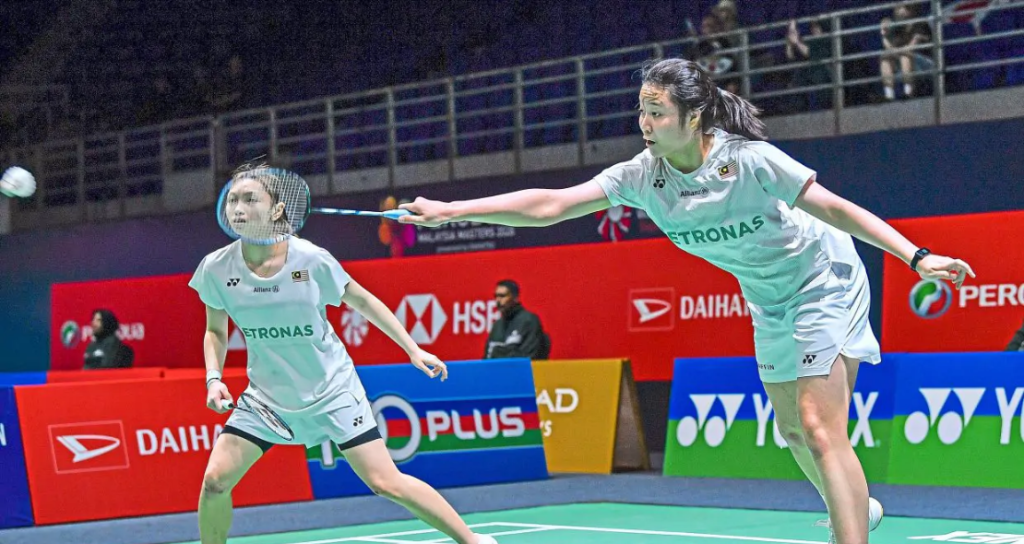 Low Zi Yu stretches to intercept the shuttlecock while partner Noraqilah Maisarah Ramdan watches alertly in their white Petronas jerseys.