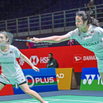 Low Zi Yu stretches to intercept the shuttlecock while partner Noraqilah Maisarah Ramdan watches alertly in their white Petronas jerseys.