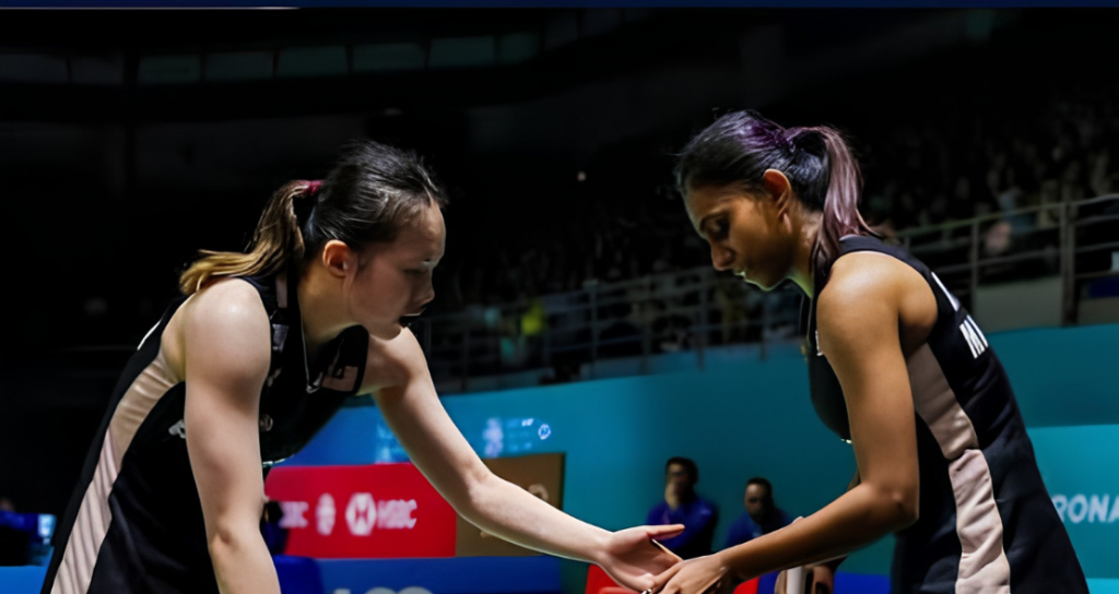 Pearly Tan and M. Thinaah touch hands on court while communicating between rallies in their black Malaysian jerseys.