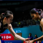 Pearly Tan and M. Thinaah touch hands on court while communicating between rallies in their black Malaysian jerseys.