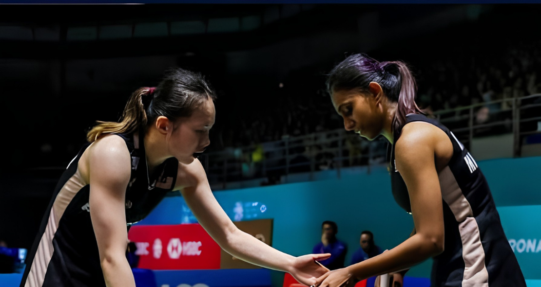 Pearly Tan and M. Thinaah touch hands on court while communicating between rallies in their black Malaysian jerseys.