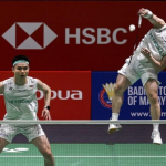 Aaron Chia leaps high in mid-air to smash while Soh Wooi Yik guards the net in their white Petronas jerseys.
