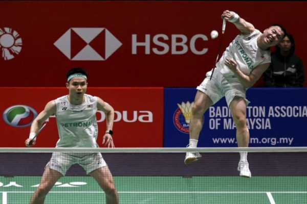Aaron Chia leaps high in mid-air to smash while Soh Wooi Yik guards the net in their white Petronas jerseys.