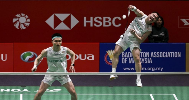 Aaron Chia leaps high in mid-air to smash while Soh Wooi Yik guards the net in their white Petronas jerseys.