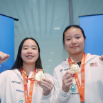 Genevie Lim and Low Zi Yu display their gold and silver medals wearing white Malaysian team jackets in front of the BAM logo.
