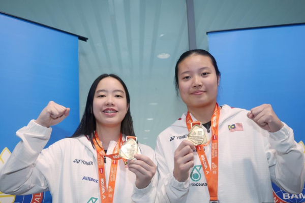Genevie Lim and Low Zi Yu display their gold and silver medals wearing white Malaysian team jackets in front of the BAM logo.