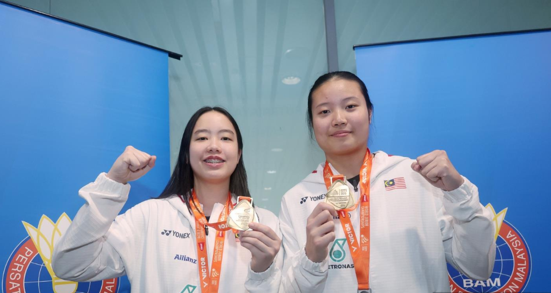 Genevie Lim and Low Zi Yu display their gold and silver medals wearing white Malaysian team jackets in front of the BAM logo.