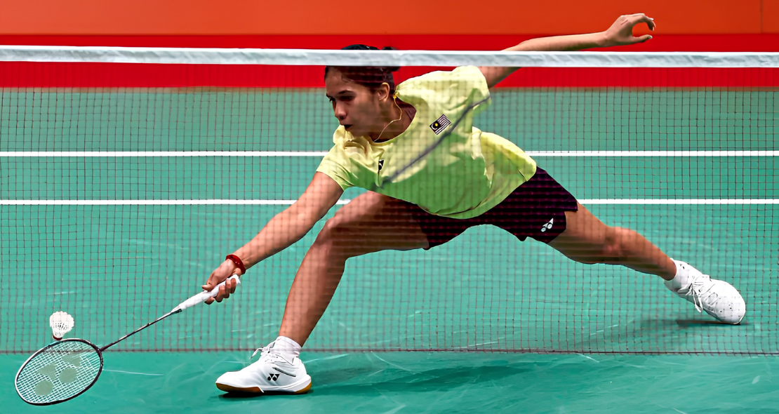 K. Letshanaa lunges deeply forward across the court to play a low net return in her yellow Malaysian national jersey.