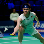 Lee Zii Jia lunges forward low to play an underhand net return in his white Petronas jersey and blue headband.
