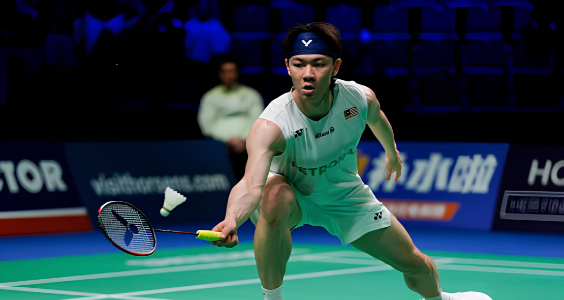 Lee Zii Jia lunges forward low to play an underhand net return in his white Petronas jersey and blue headband.