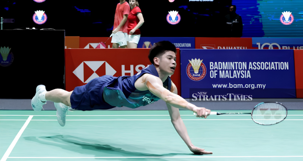Leong Jun Hao executes a full horizontal dive across the court to retrieve the shuttlecock in his blue Petronas jersey.
