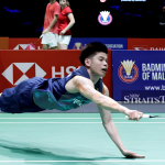 Leong Jun Hao executes a full horizontal dive across the court to retrieve the shuttlecock in his blue Petronas jersey.